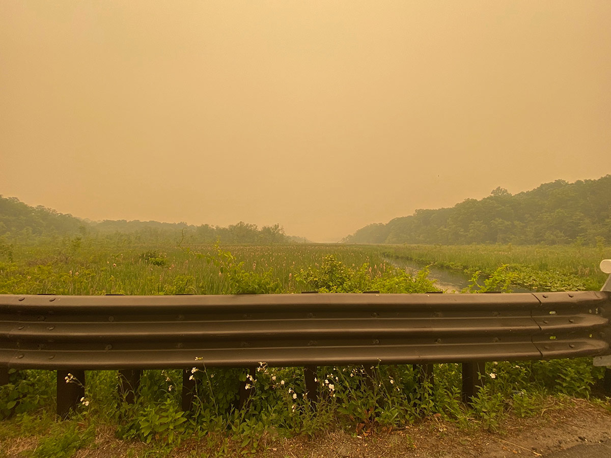 A roadside overlooking a field and stream. The entire view is obscured by orange haze, the result of wildfire smoke from a series of wildfires in 2023.