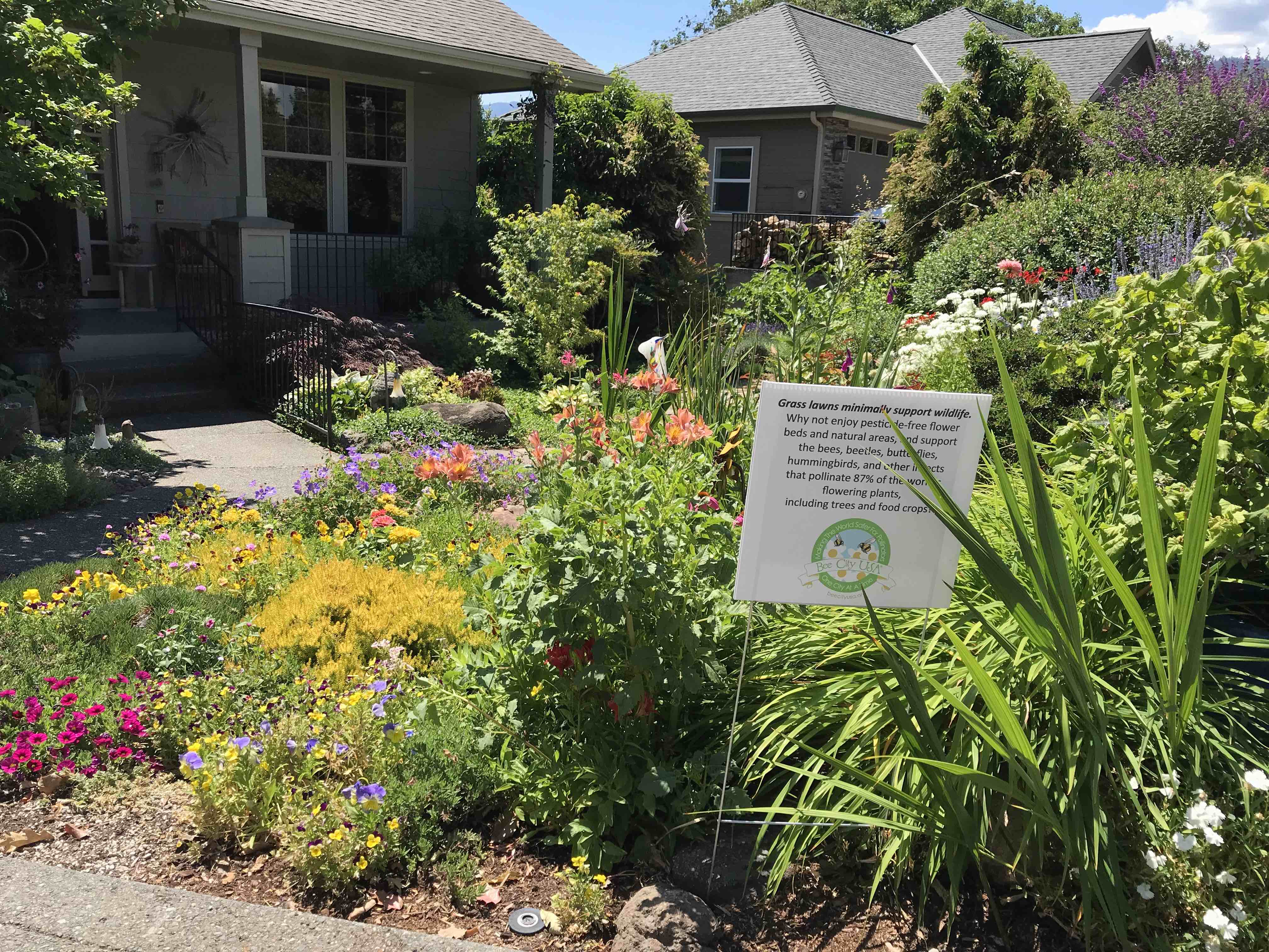 A colorful garden with plants of various heights and flower colors has a sign in front with the Bee City USA's green, circular logo with bees in the middle.
