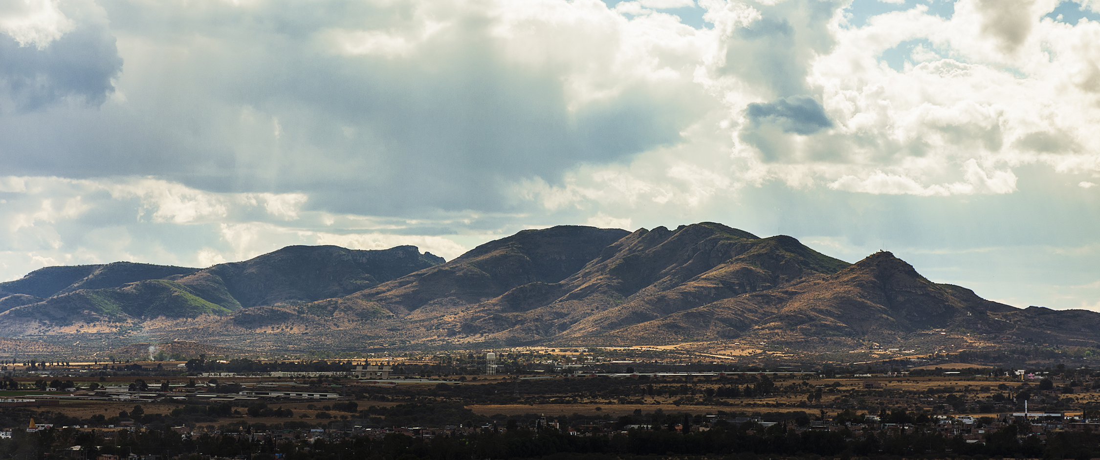 A photo of the landscape of central Mexico. The land in the foreground is flat. Behind, a range of mountains rises toward the clouds.