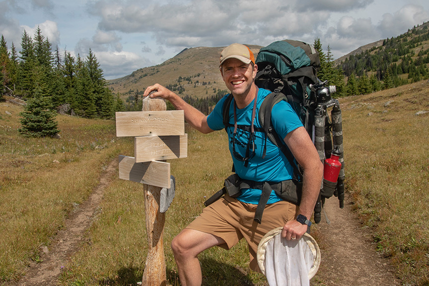 The author grins as he poses by a wooden trail sign, with a mountain in the background. He is carrying a big backpack and holding a net.