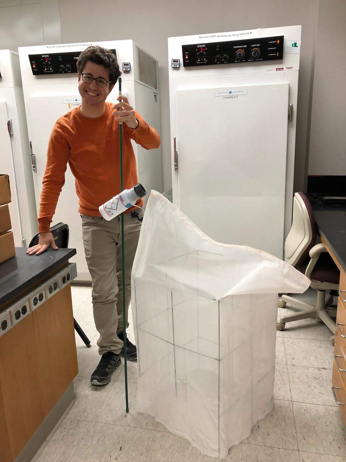A smiling young man stands in a lab, next to a white mesh box. He is holding a pole with a plastic container attached to it.