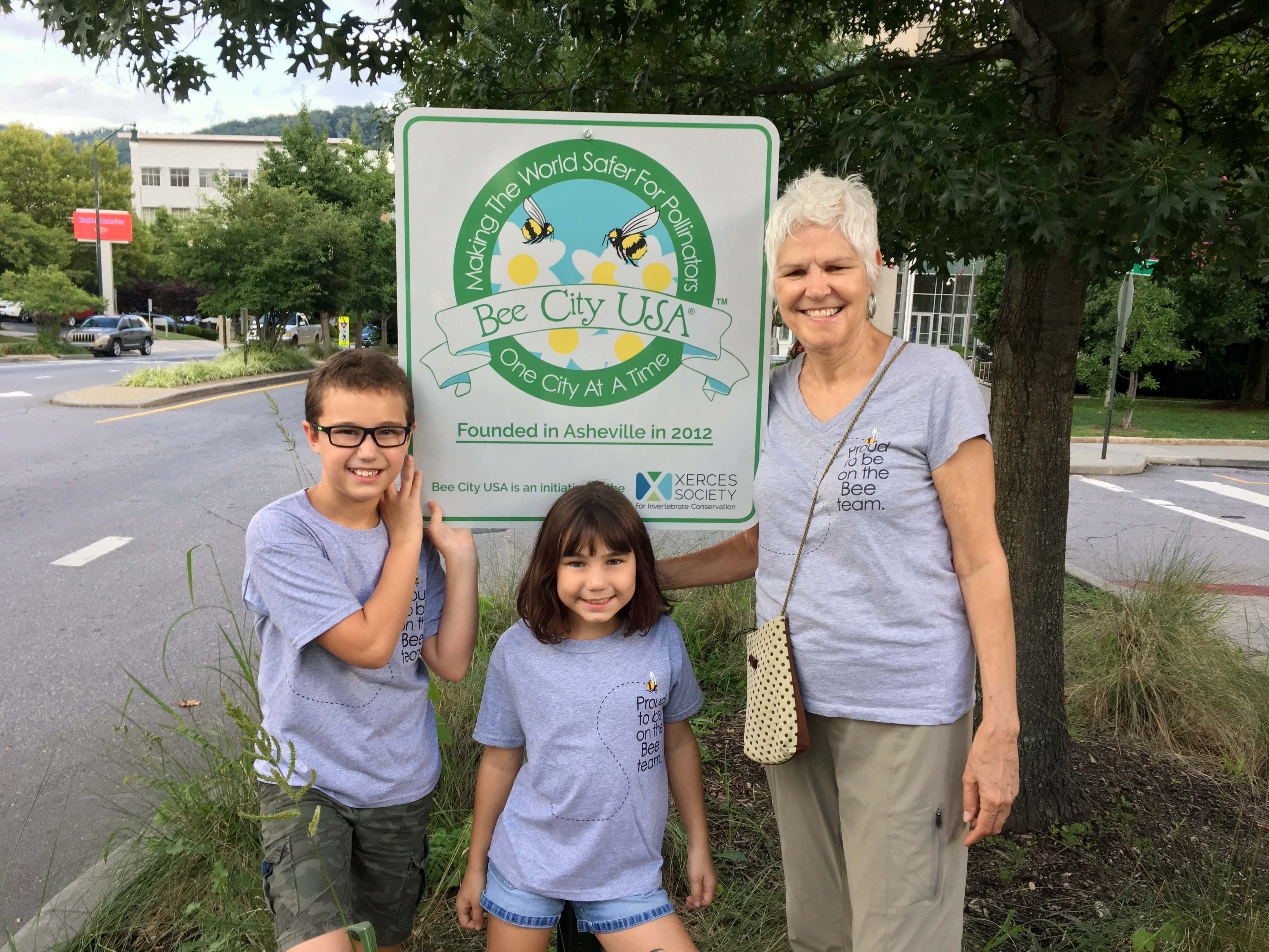 A tall woman with short, white hair poses with two children by a sign with the Bee City USA logo, that reads "founded in Asheville in 2012."
