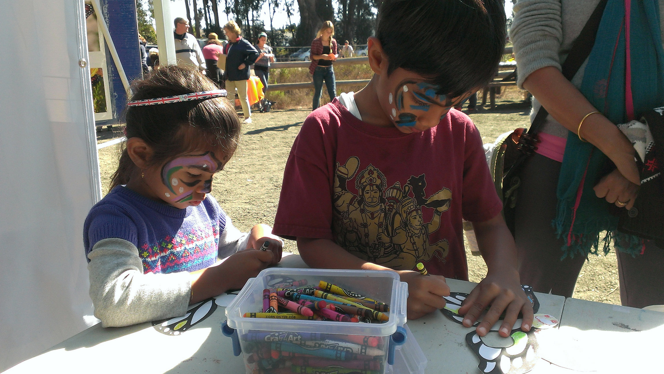 Two children, their faces decorated with face paints, concentrate on coloring monarch butterfly drawings