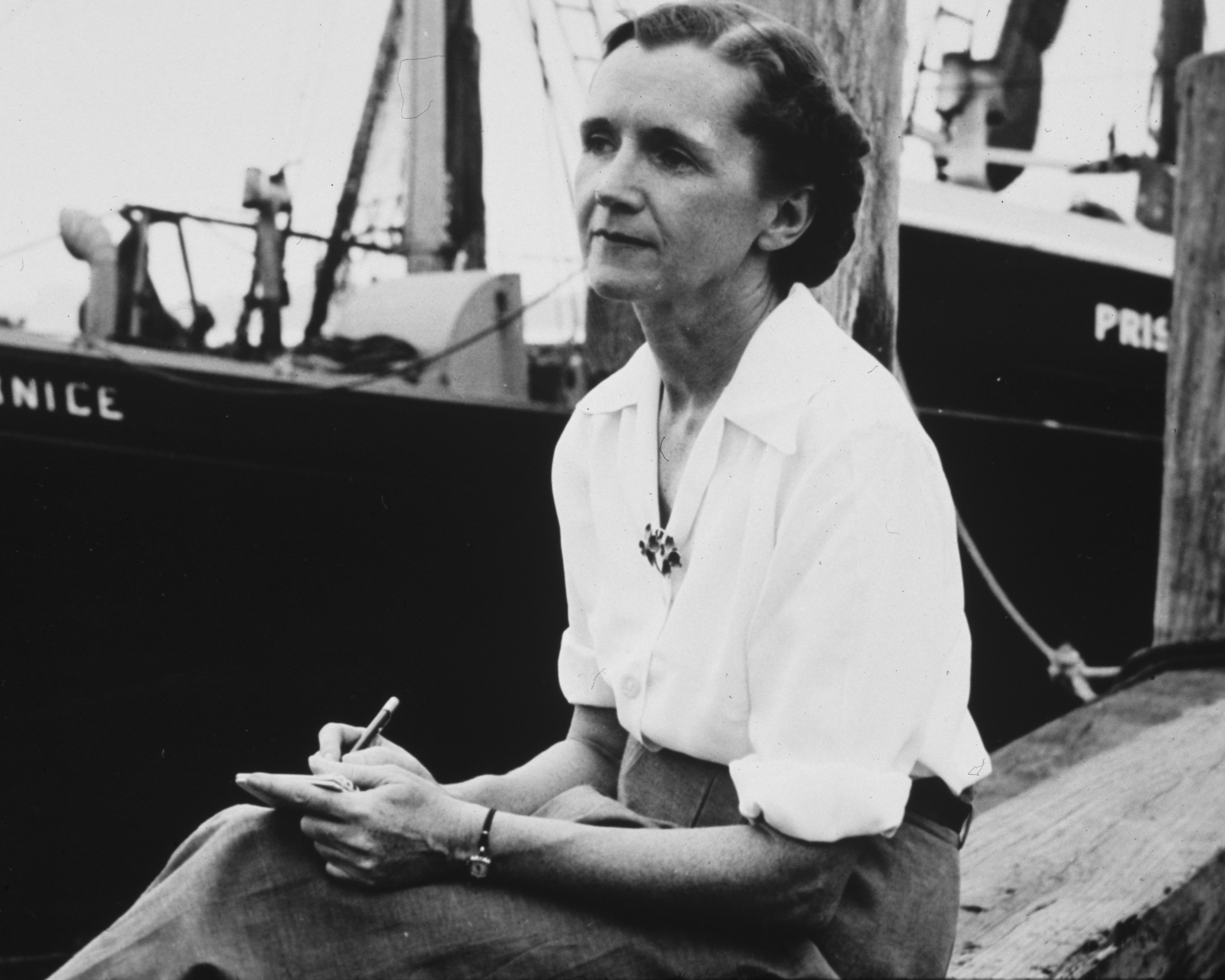 Rachel Carson sits with pen in hand on a dock, with a boat behind her, in this black-and-white historic photo.