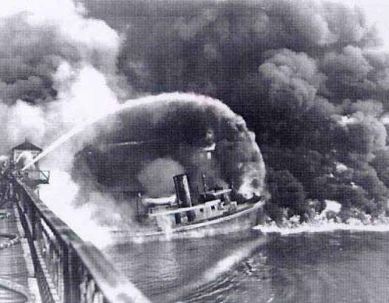 A firefighter sprays water from a bridge onto the burning Cuyahoga River in this historical photo, in black and white..