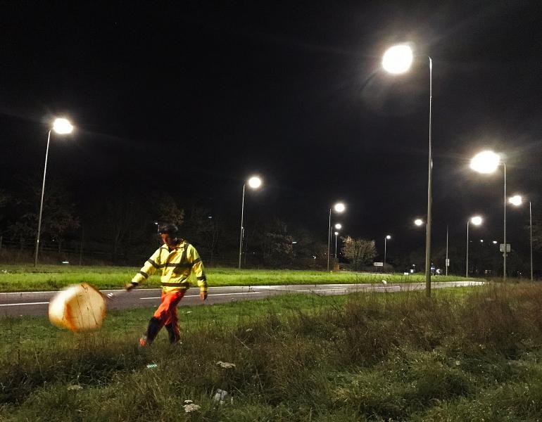 A person wearing fluorescent clothing and a headlamp swings a net alongside a roadway lit by streetlamps at night.