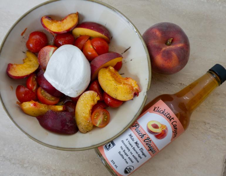 A bottle of vinegar lies on a table beside a bowl full of slices of peach and cherry tomatoes