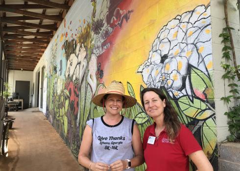 Two women in grey and red shirts stand in front of a colorful mural that shows flowers and insects at the State Botanical Garden of Georgia