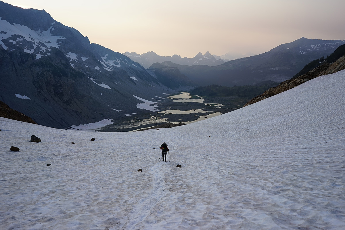 A Xerces conservation biologist hikes down from a small glacier that is now mostly just a snowfield. A chain of meltwater lakes can be seen in the distance, with smoke from nearby wildfires on the horizon.