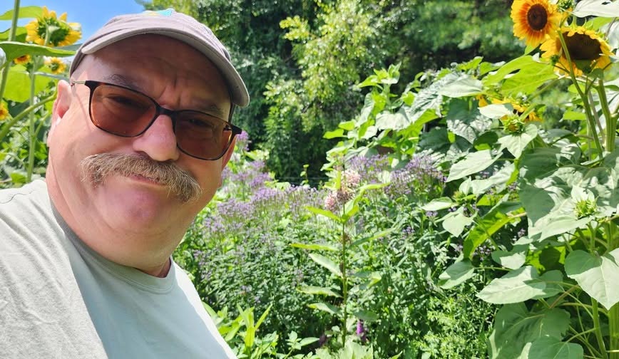 A man stands smiling in front of tall flowering plants like sunflowers