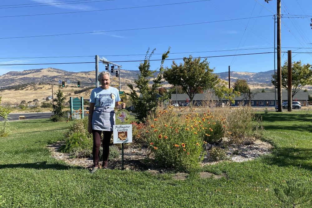 A woman with short, gray hair stands before blooming flowers and a pollinator habitat sign. Blue sky, power lines and hills are in the background.