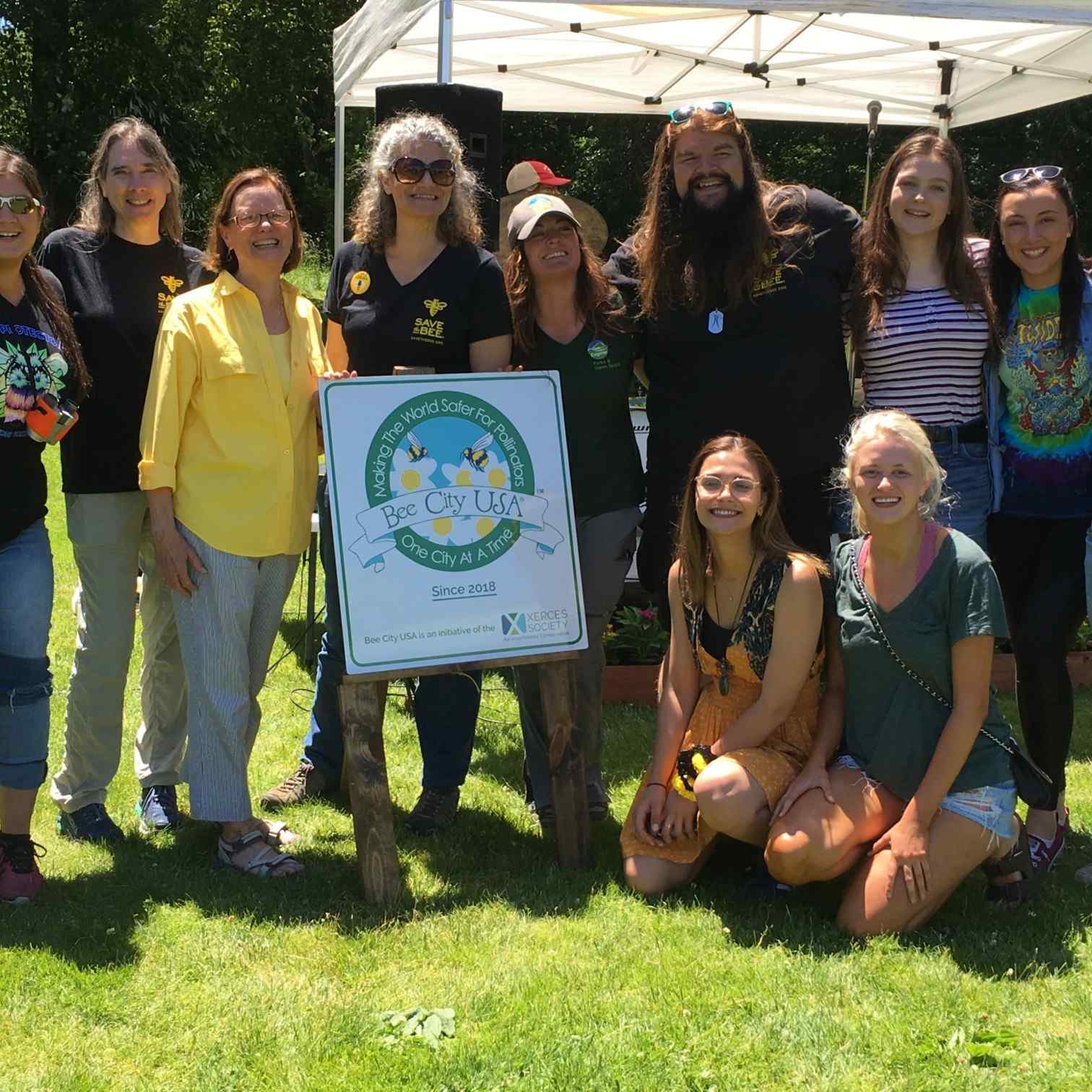 A group of people pose outdoors with a “Bee City USA” sign at a community event.