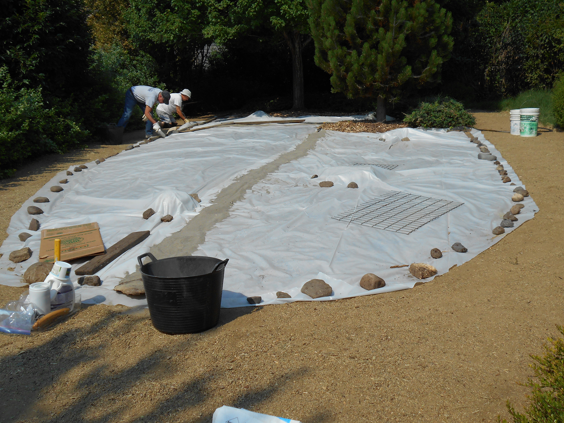 Two people work to secure the edge of a large oval of plastic sheet to hold it in place over an area of weeds. The weeds will wilt under the plastic, leaving the area clear and ready for a pollinator garden