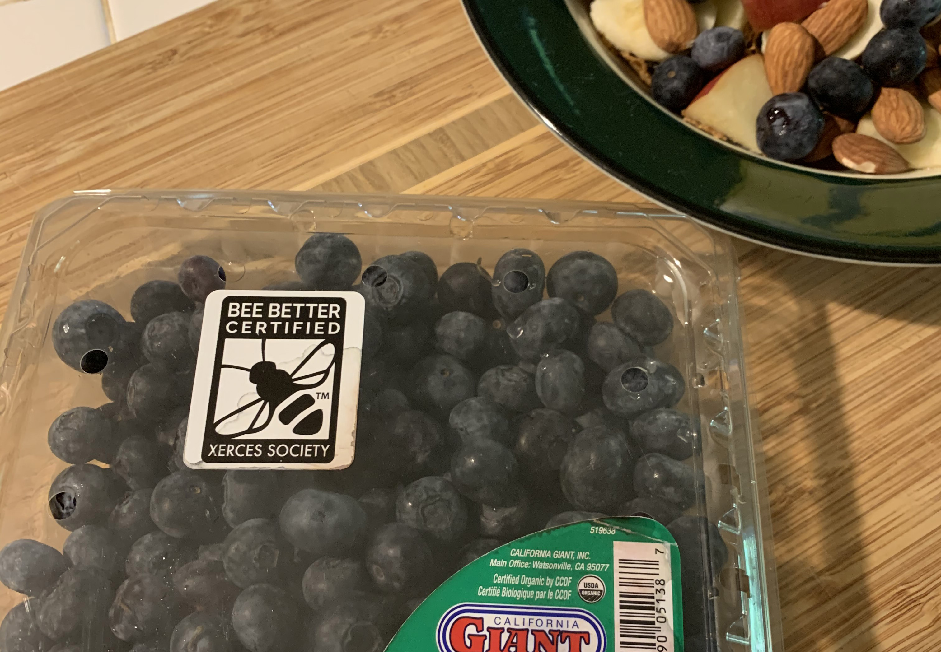 A clear plastic container full of round, dark colored blueberries. The container has a green label with white writing in the lower right corner and in the top right corner, a black-and-white label that says the blueberries are Bee Better Certified. Beside the container is a green cereal bowl containing blueberries, almonds, and sliced apples.