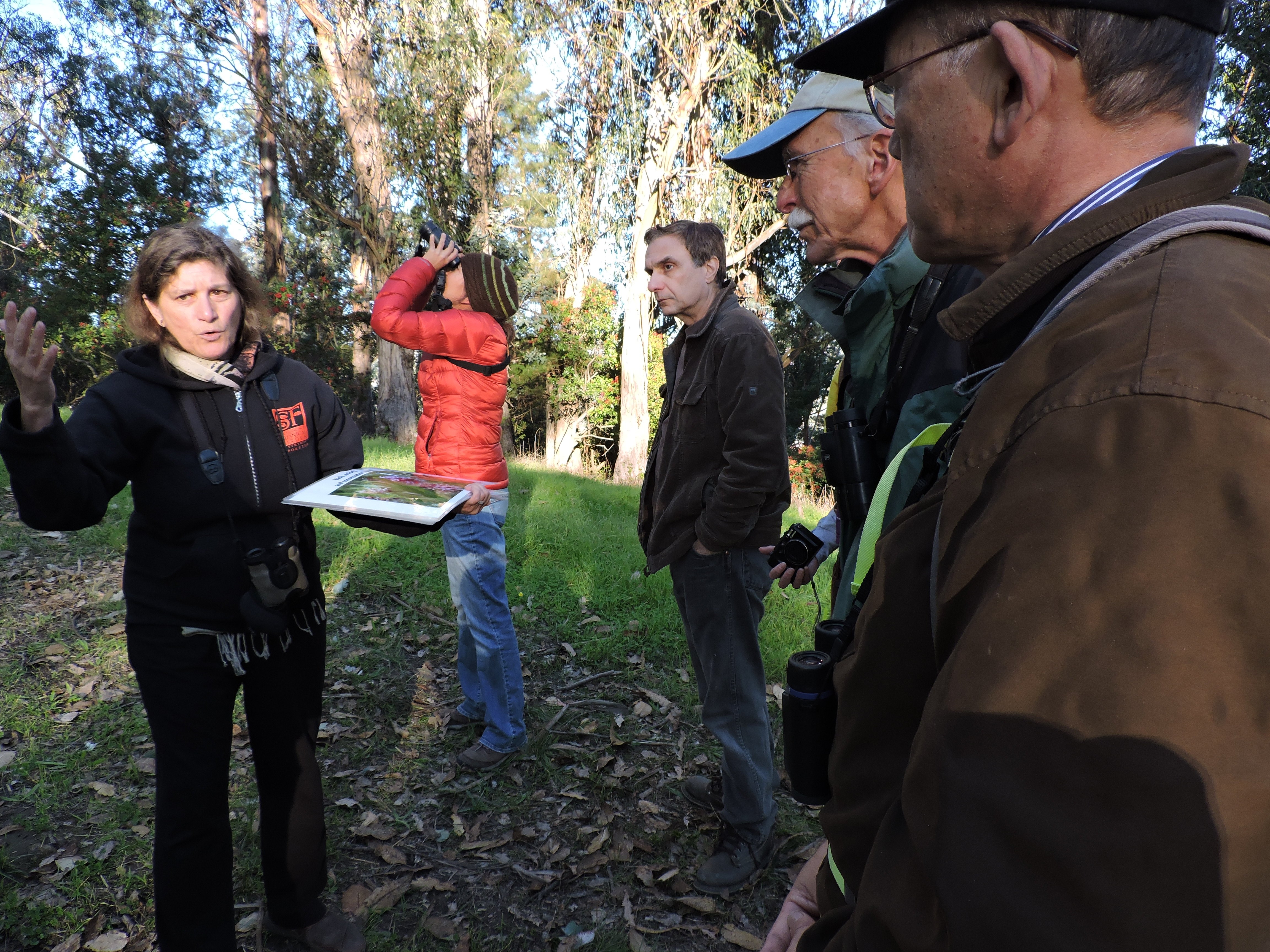 A woman in a dark coat (left) speaks to an assembled group of volunteers (right) in a wooded setting.