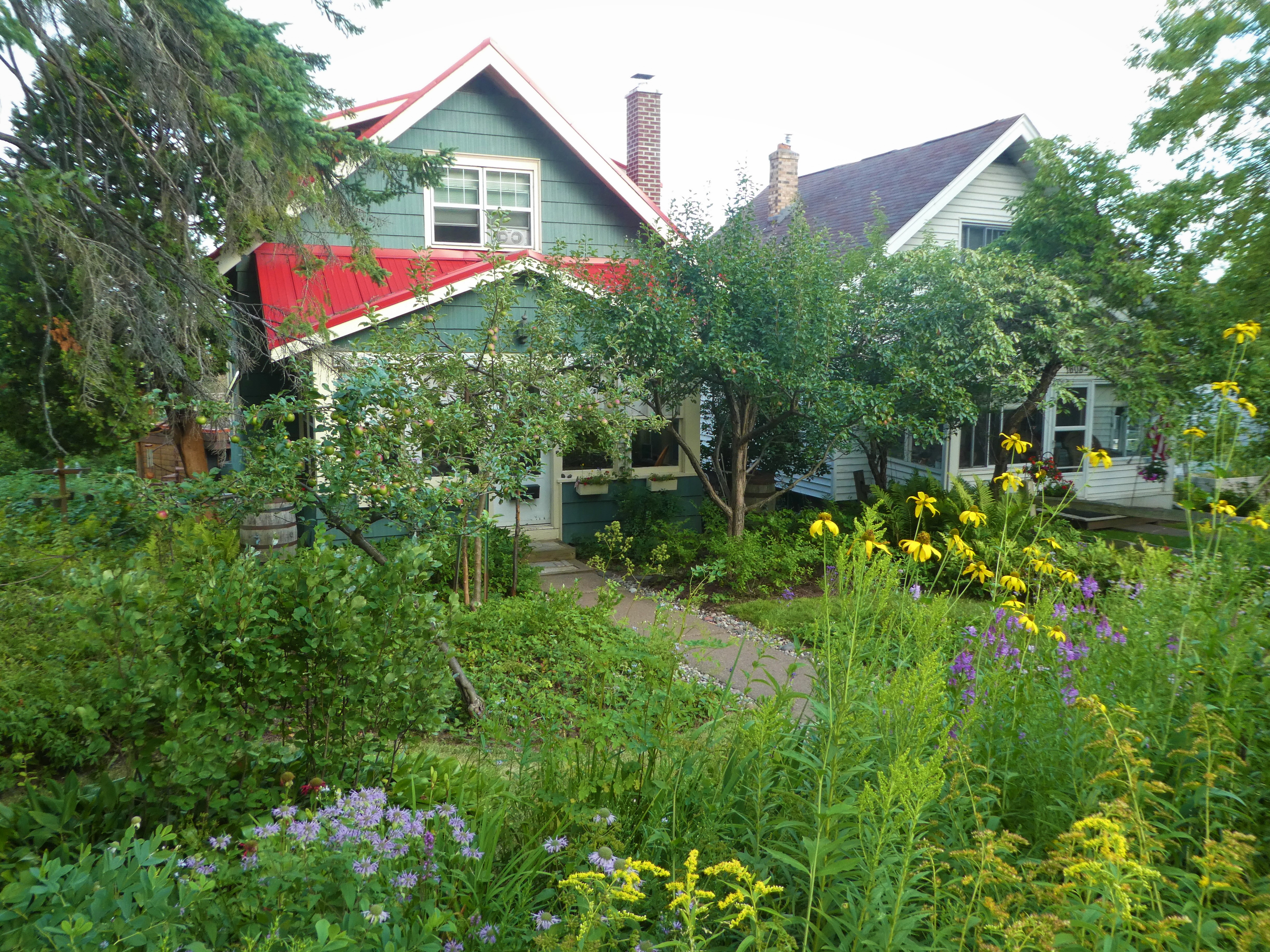 A green house is nearly obscured by a lush, green yard bursting with colorful flowers in the foreground, and fruit trees nearer to the house.
