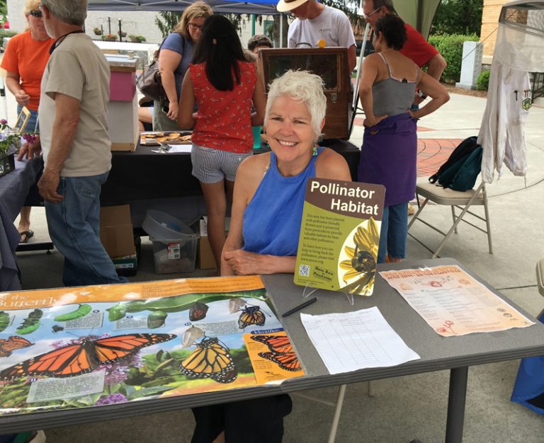 Phyllis Stiles, Bee City USA founder, tables at an event. She is seated at a table with a lot of materials, including a Xerces Society pollinator habitat sign.