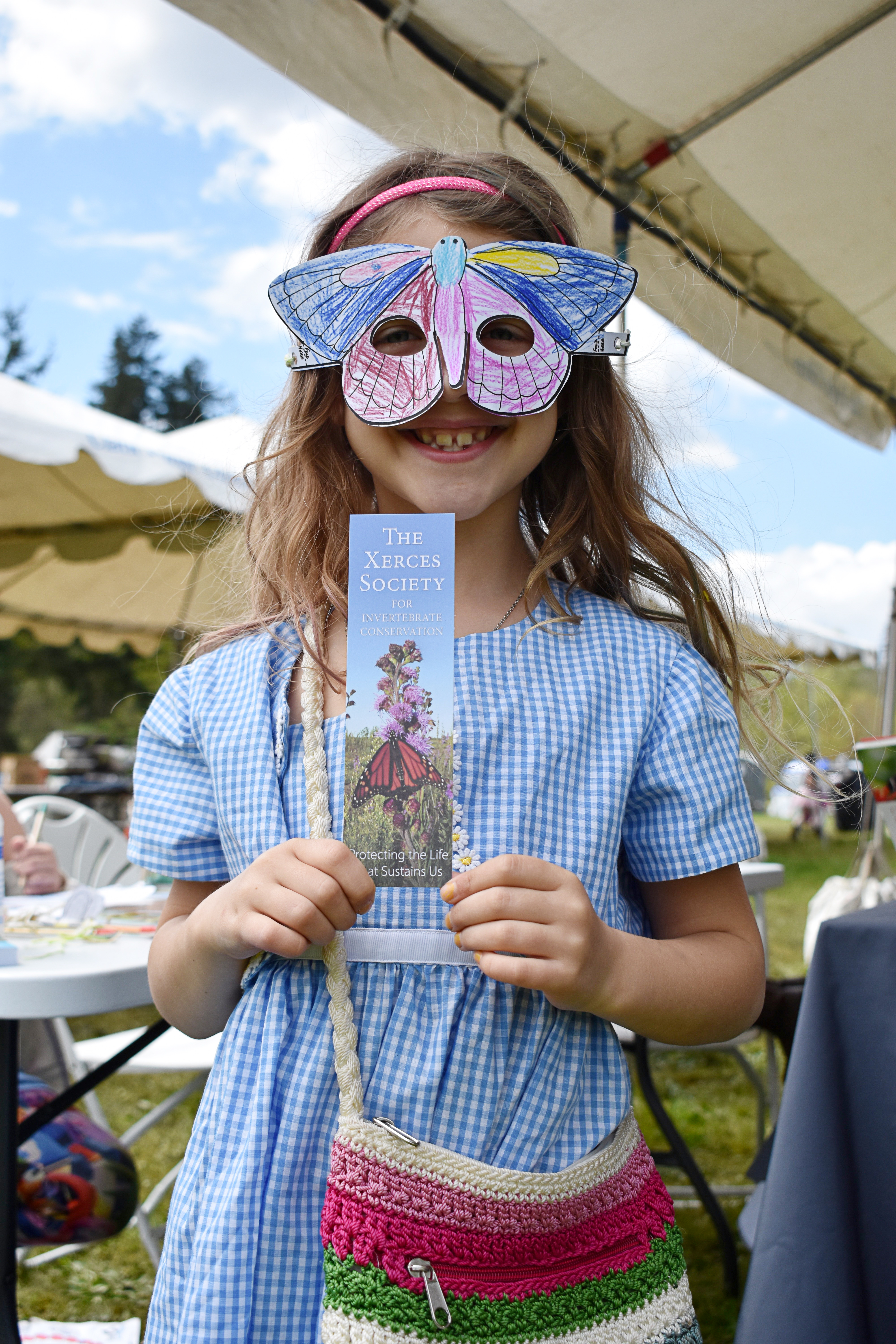 A girl smiles as she wears a hand-colored paper butterfly mask and holds up a Xerces Society bookmark with a photo of a monarch butterfly (orange and black wings) on a purple flower in a meadow.