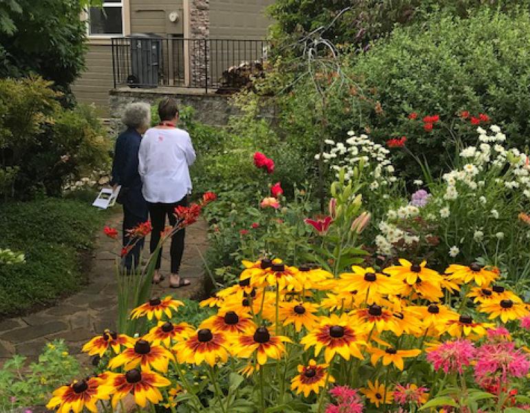Two women walk through a garden with abundant, colorful flowers.