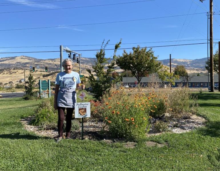 A woman with short, gray hair stands smiling in front of blooming native flowers and a pollinator habitat sign. Blue sky, power lines and hills are in the background.