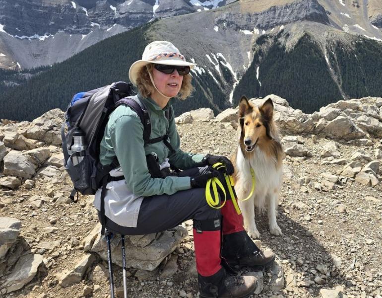 woman in hiking gear sits near her collie dog on top of a mountain summit