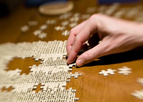 Pieces of a puzzle are being assembled on a dark, wooden table. A hand moves the pieces.