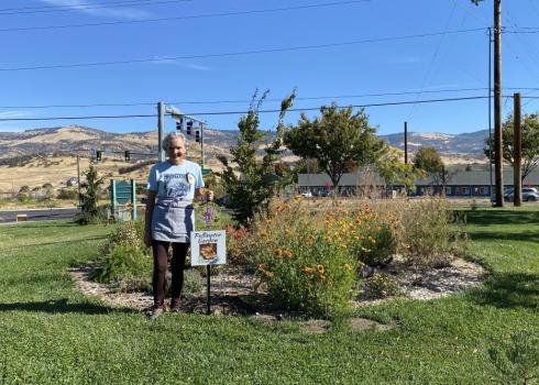 A woman with short, gray hair stands smiling in front of blooming native flowers and a pollinator habitat sign. Blue sky, power lines and hills are in the background.