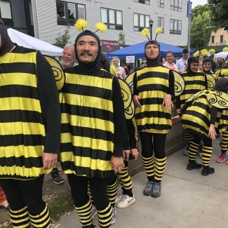 A long line of people of diverse ages, genders, and races are all sporting bright yellow and black-striped bee outfits in an outdoor festival setting.