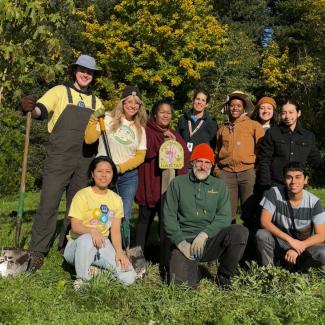 10 people in work clothes with shovels gather around a pollinator habitat sign in a garden.