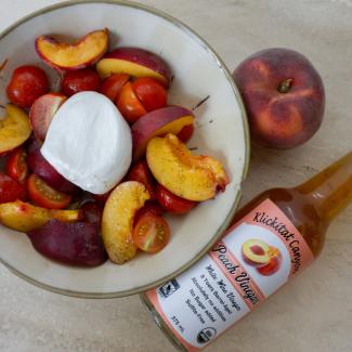 A bottle of vinegar lies on a table beside a bowl full of slices of peach and cherry tomatoes