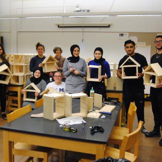 A group of young people standing a classroom holding wooden insect houses that they have been building. Some people have blonde hair, some brown, and some black. A few of the group are wearing head scarfs. The insect houses are square frames with a pointed roof.