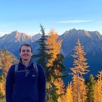 A person wearing a blue fleece and backpack stands among golden autumn trees with rugged mountains and a clear blue sky in the background