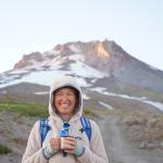 Smiling person in a hooded fleece holding a mug on a rocky mountain trail, with a snow-covered peak in the background